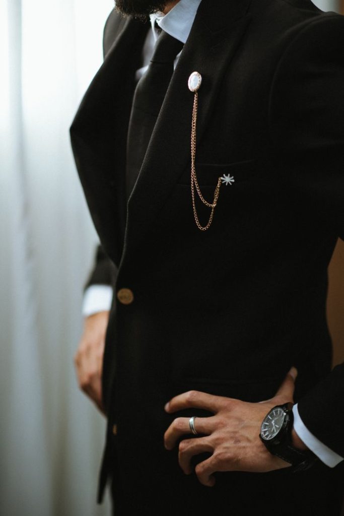 A man in a formal black suit with gold chain detail and wristwatch, exuding elegance.